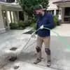 A plumber with a beard, wearing knee pads and a blue uniform, operating a hydro-jetting hose to clear a drain on a paved driveway in front of a house.