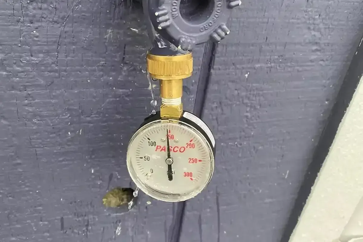 A water pressure gauge attached to an outdoor spigot, indicating a pressure reading, with water droplets visible around the connection point.