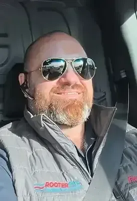 A smiling, bearded man wearing sunglasses, an earpiece, and a grey RooterPAL branded vest, sitting in a vehicle.