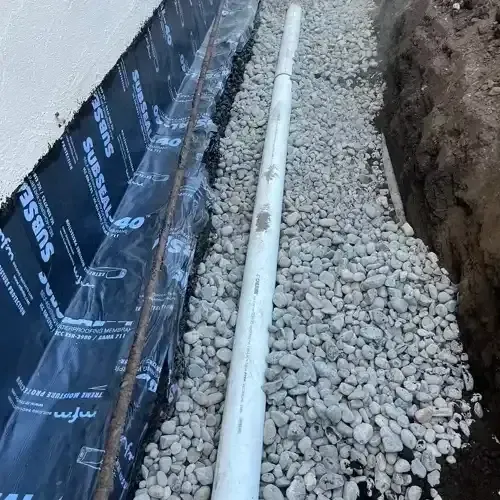A newly installed drainage system for foundation waterproofing, showing a white pipe laid in gravel next to a black Subseal 40 membrane.